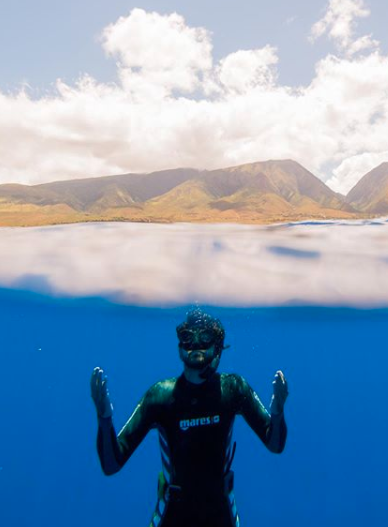 a half-under, half-above water shot of a man snorkeling, with mountains in the background